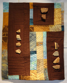 Quilt in browns and golds with bits of pottery on the surface